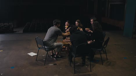 Lunch-of-young-actors-in-black-suits-eating-pizza-during-preparation-for-the-performance-together-with-their-director-a-brunette-man-in-a-gray-T-shirt-on-the-stage-of-a-modern-theater