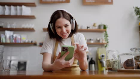 Happy-brunette-girl-waiter-in-a-yellow-apron-wearing-white-wireless-headphones-listens-to-music-and-watches-a-video-during-a-break-at-work-in-a-cafe