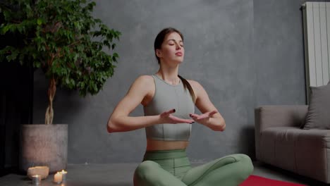 Zoom-in-Confident-brunette-girl-in-a-gray-top-and-green-sweatpants-does-yoga-sits-in-the-lotus-position-and-practices-breathing-practices-in-a-modern-apartment-at-home-near-a-houseplant