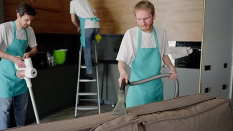 Zoom-Out-portrait-of-a-confident-guy-a-blond-cleaner-in-a-white-T-shirt-and-a-blue-apron-who-is-cleaning-the-sofa-with-a-vacuum-cleaner-together-with-his-colleagues-cleaning-the-kitchen-on-call-from-a-cleaning-company