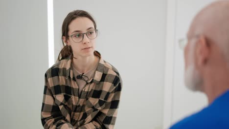 A-young-brunette-girl-with-glasses-and-a-plaid-shirt-interviews-a-confident-and-experienced-male-doctor-in-a-blue-uniform-in-a-modern-clinic.-Over-his-shoulder,-a-confident-man-doctor-talks-to-a-young-girl-interviewer-and-talks-about-the-doctors-profession