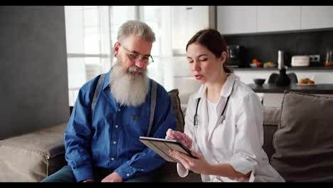 Una-Doctora-Morena-Con-Bata-Blanca-Sostiene-Una-Tableta-Electrónica-En-Sus-Manos-Y-Le-Muestra-A-Un-Hombre-Mayor-Con-Gafas-Y-Una-Exuberante-Barba-Gris-Lo-Que-Debe-Hacer-Para-Mantener-Su-Salud-En-Un-Apartamento-Moderno.