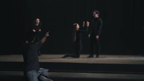 Rear-view-of-a-confident-male-director-with-papers-in-his-hands-waving-his-arms-and-gesticulating-showing-the-actors-how-to-play-correctly-during-a-rehearsal-of-actors-in-black-suits-on-stage-in-the-theater