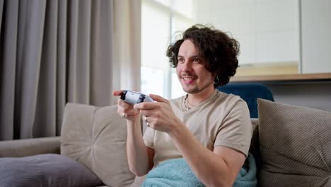 Happy-brunette-guy-with-curly-hair-and-mustache-plays-video-games-and-wins-with-a-joystick-and-sits-on-the-sofa-at-home-in-a-modern-apartment