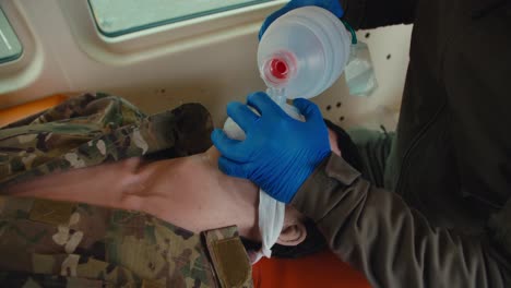 Close-up-of-a-confident-male-army-medic-in-a-dark-green-uniform-and-blue-gloves-doing-artificial-respiration-to-an-unconscious-man-using-a-matte-white-special-bottle-for-artificial-respiration-in-the-ambulance-cabin