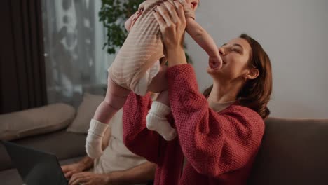 Happy-young-brunette-mother-in-a-red-sweater-lifts-and-plays-with-her-little-baby-daughter-while-her-husband-works-using-a-laptop-while-sitting-on-a-gray-sofa-in-a-modern-apartment