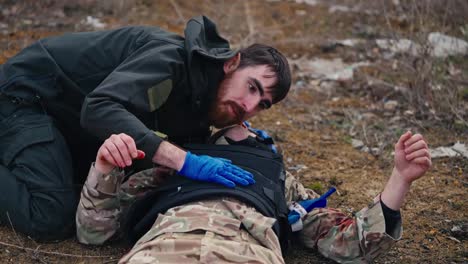 Close-up-of-a-confident-male-military-brunette-with-a-beard-in-a-dark-green-military-uniform-listens-to-the-breathing-and-holds-the-chest-of-a-wounded-soldier-during-combat-and-providing-first-aid-outside-the-city