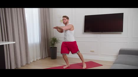 Confident-blond-man-with-stubble-in-a-white-T-shirt-and-red-shorts-does-morning-exercises-and-does-leg-and-arm-crunches-while-standing-on-the-Red-carpet-in-a-modern-apartment-at-home