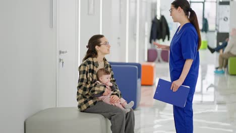 A-young-brunette-girl-the-mother-of-a-little-baby-girl-sits-in-the-corridor-of-a-modern-clinic-and-waits-for-her-turn-in-the-office-of-a-pediatrician-doctor.-While-a-brunette-girl-doctor-in-a-blue-uniform-comes-up-and-reassures-the-young-mother