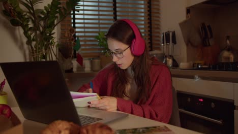 A-confident-brunette-girl-in-red-wireless-headphones-and-a-sweater-studies-foreign-languages-taking-notes-from-a-lesson-in-her-notebook-while-sitting-in-the-kitchen-in-a-modern-apartment-near-the-flag-of-Italy