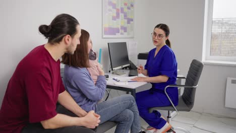 Young-happy-family-a-brunette-guy-in-a-red-T-shirt-together-with-his-wife-and-little-daughter-take-advice-from-a-childrens-doctor-and-a-girl-pediatrician-in-a-blue-uniform-and-glasses-in-a-modern-clinic