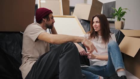 A-brunette-girl-in-a-white-T-shirt-communicates-with-her-boyfriend-in-a-red-hat-with-stubble-while-they-sit-near-a-sofa-covered-with-black-polyethylene-among-a-large-number-of-boxes-and-a-house-plant-in-a-modern