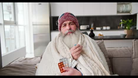 Portrait-of-a-sick-elderly-man-with-a-gray-lush-beard-in-a-red-hat-and-a-white-plaid-holding-a-thermometer-and-a-set-of-pills-while-sitting-on-a-modern-sofa-in-an-apartment