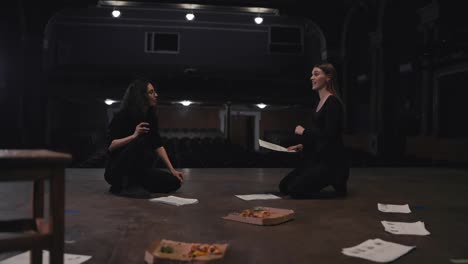 A-confident-girl-with-brown-hair-and-a-black-suit-holds-sheets-of-paper-with-a-script-in-her-hands-and-expressively-addresses-her-fellow-actress-during-her-rehearsal-for-a-play-on-the-theater-stage