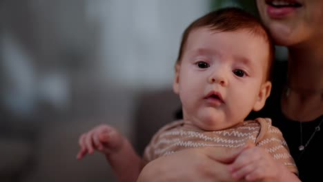 Retrato-En-Primer-Plano-De-Una-Niña-Pequeña-Con-Una-Chaqueta-Marrón-Claro-Sentada-En-Los-Brazos-De-Su-Madre-Y-Mirando-A-Su-Alrededor-En-Un-Apartamento-Moderno.