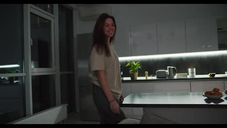 A-happy-brunette-girl-in-a-beige-T-shirt-approaches-her-husband-from-behind-while-he-in-a-black-jacket-and-beige-shorts-works-sitting-on-the-sofa-behind-a-gray-laptop-in-the-evening-in-a-modern-studio-apartment