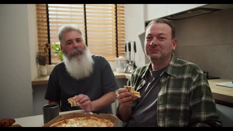 Portrait-of-a-happy-LGBT-couple-of-two-men-brunette-in-a-checkered-shirt-and-an-older-man-with-gray-hair-and-a-lush-gray-beard-who-eat-pizza-in-the-kitchen-during-their-lunch-and-are-happy