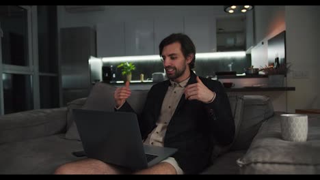 A-happy-brunette-man-with-stubble-in-a-black-jacket-and-beige-shorts-sits-on-the-sofa-and-communicates-via-video-conference-using-a-laptop-while-his-brunette-girlfriend-relaxes-in-the-kitchen-in-a-modern-studio-apartment