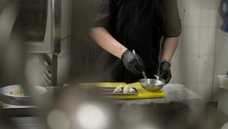 Filmado-Desde-Un-Costado,-Un-Chef-Profesional-Con-Uniforme-Negro-Y-Guantes-De-Goma-Rellena-Pequeños-Trozos-De-Productos-De-Harina-Y-Prepara-Un-Plato-En-La-Cocina-De-Un-Restaurante.