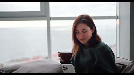 A-happy-brunette-woman-in-a-dark-green-jacket-drinks-tea-from-a-brown-cup-and-looks-at-a-white-phone-during-her-work-and-relaxation-at-home-in-a-modern-apartment-overlooking-the-sea