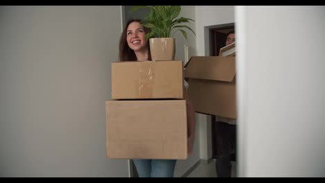 A-happy-brunette-girl-together-with-her-boyfriend-enters-a-new-apartment-holding-large-wooden-boxes-and-house-plants-in-her-hands-in-her-new-purchased-apartment