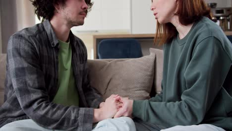 Zoom-Out-a-brunette-girl-in-a-green-sweater-talks-about-a-serious-topic-about-relationships-with-her-boyfriend-a-brunette-with-curly-hair-in-a-checkered-shirt-sitting-on-the-sofa-in-a-modern-apartment-at-home