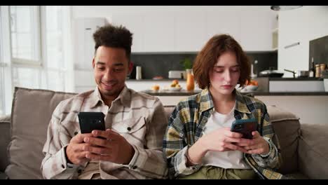 Calm-couple-a-cheerful-man-a-young-brunette-with-Black-skin-color-in-a-cream-plaid-shirt-scrolling-social-networks-while-sitting-on-the-sofa-with-his-girlfriend-who-is-also-looking-at-something-on-her-smartphone-in-a-modern-studio-apartment