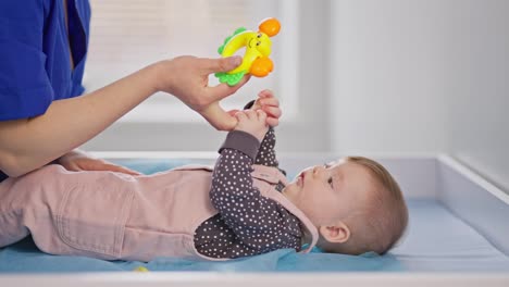 Side-view-of-a-little-girl-baby-in-a-pink-jumpsuit-lies-on-a-bed-in-a-pediatrician-doctors-office-who-plays-with-him-using-a-childs-toy-in-a-modern-clinic-for-children