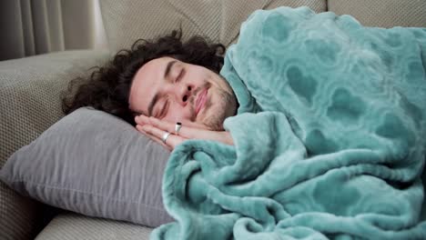 Turn-on-the-guy-with-curly-hair-and-a-mustache-lies-on-a-gray-pillow,-wrapped-in-a-blue-blanket-and-sleeps-on-the-sofa-at-home