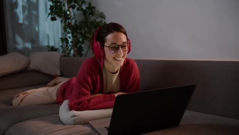 Happy-young-brunette-girl-with-glasses-in-red-wireless-headphones-and-a-sweater-lies-on-a-gray-sofa-and-studies-foreign-languages-with-the-help-of-online-teachers-and-lessons-in-a-modern-apartment