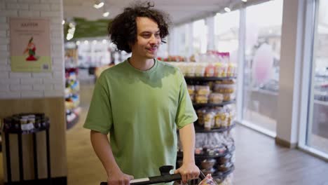 Happy-brunette-guy-with-curly-hair-and-mustache-walks-and-looks-at-goods-in-a-grocery-store-while-shopping