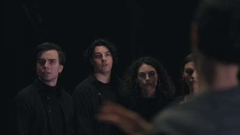 Close-up,-four-confident-young-actors-in-black-suits-stand-in-a-row-and-listen-to-their-director-Skip-and-papers-and-the-plot-on-them-during-a-rehearsal-for-a-play-in-the-theater