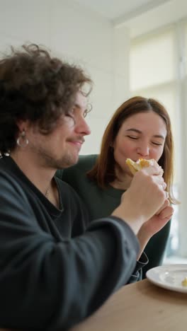 Vertical-video-of-a-happy-brunette-girl-biting-a-sandwich-that-her-boyfriend-feeds-her-during-breakfast-in-the-morning