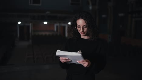 Portrait-of-a-confident-brunette-girl-with-curly-hair-in-glasses-in-a-black-suit-the-actress-holds-a-script-in-her-hands-and-recounts-the-text-of-her-character-in-the-theater-during-a-rehearsal