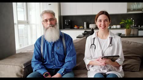 Retrato-De-Una-Doctora-Feliz-Con-Una-Bata-Blanca-Con-Un-Estetoscopio-Y-Una-Tableta-En-Sus-Manos-Que-Está-Sentada-En-Un-Sofá-Junto-Con-Un-Hombre-Mayor-Con-Una-Exuberante-Barba-Gris-Con-Una-Camisa-Azul-En-Un-Apartamento-Moderno-Durante-Un-Examen-Domiciliario.