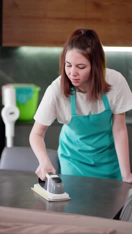 Vertical-video-of-a-confident-brunette-cleaning-lady-in-a-blue-apron-polishing-a-glossy-table-in-a-modern-apartment.-Portrait-of-a-confident-brunette-girl-a-professional-cleaner-who-does-cleaning-on-call