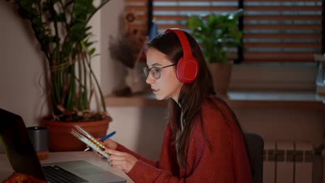 A-confident-brunette-girl-in-wireless-headphones-and-glasses-in-a-modern-apartment-in-the-kitchen-studies-foreign-languages-and-takes-notes-on-the-rules-of-pronunciation