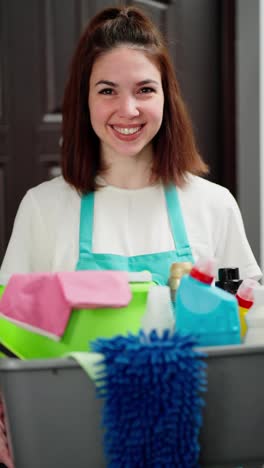 Vertical-video-portrait-of-a-happy-girl-professional-cleaner-in-blue-aprons-with-many-cleaning-items-in-her-hands-in-the-house