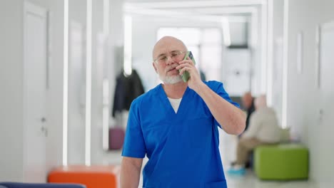 A-confident-elderly-man-with-a-beard-a-doctor-walks-along-the-corridor-of-a-bright-clinic-and-talks-on-the-phone-during-the-working-day.-A-confident-elderly-doctor-walks-through-the-clinic-talking