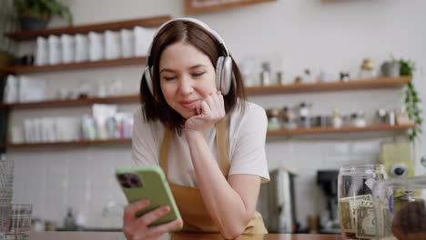 Happy-and-cheerful-brunette-waiter-girl-in-a-yellow-apron-looks-at-social-networks-and-listens-to-music-using-white-wireless-headphones-and-a-green-smartphone-standing-at-the-counter-in-a-cafe