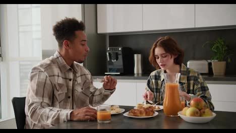 A-young-adult-girl-with-brown-hair-and-a-bob-hairstyle-in-a-green-checkered-shirt-communicates-with-her-young-boyfriend-with-Black-brunette-skin-color-at-a-black-kitchen-table-in-the-kitchen-in-a-modern-apartment