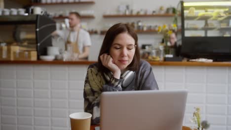 Porträt-Einer-Brünetten-Frau-Mit-Kabellosen-Kopfhörern,-Die-An-Einem-Tisch-Vor-Einem-Laptop-In-Einem-Café-Sitzt-Und-Während-Ihres-Arbeitstages-Posiert