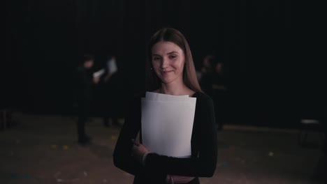 Portrait-of-a-confident-young-girl-actor-in-a-black-suit-who-crosses-her-arms-on-her-chest-while-holding-play-script-papers-in-her-hands-during-a-rehearsal-in-the-theater