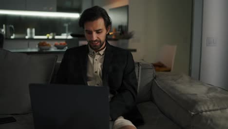 Confident-brunette-man-with-stubble-in-a-black-jacket-and-beige-shirt-and-shorts-sits-on-a-gray-sofa-and-works-on-his-laptop-in-a-modern-studio-apartment-leaning-against-his-wife-and-having-a-good-time-checking-the-time-while-working-remotely