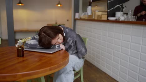 A-tired-brunette-girl-in-a-gray-T-shirt-and-white-wireless-headphones-lies-and-relaxes-on-a-laptop-while-sitting-at-a-table-in-a-cafe