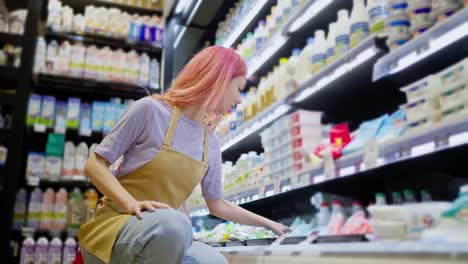 Side-view-of-a-confident-girl-with-pink-hair-working-in-a-supermarket-and-placing-goods-on-the-dairy-products-counter-in-a-supermarket