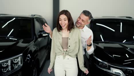 Happy-middle-aged-man-opens-his-girlfriend-eyes-with-his-brunette-hands-while-informing-her-about-buying-a-modern-car-at-a-car-dealership.-Happy-brunette-girl-receives-a-surprise-while-buying-a-car-at-a-car-dealership