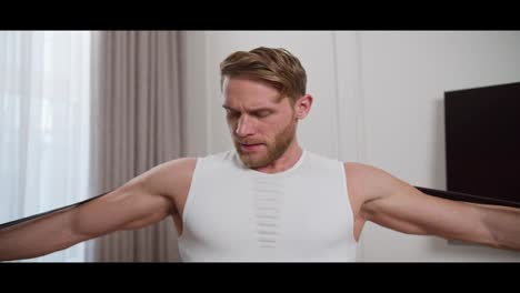 Straight-view-of-a-blond-athletic-man-with-stubble-a-man-in-a-white-T-shirt-does-morning-exercises-and-swings-his-arms-using-a-special-rubber-expander-at-home-in-a-modern-bright-apartment-at-home