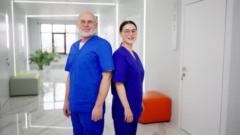 Confident-Back-to-back-Duo-of-an-experienced-elderly-man-doctor-in-a-blue-uniform-together-with-his-assistant-brunette-girl-with-glasses-doctor-in-a-modern-clinic-folding-their-arms-on-their-chest-as-a-sign-of-confidence