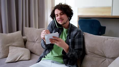 Confident-business-guy-brunette-with-curly-hair-communicates-on-the-phone-and-writes-down-important-things-in-a-conversation-on-a-notebook-at-home-on-the-sofa-in-a-modern-apartment